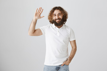 Fototapeta premium Hi, my friend. Portrait of good-looking ordinary eastern male with curly hair and beard raising hand and waving in greeting gesture, welcoming familiar person on street or giving five to mate