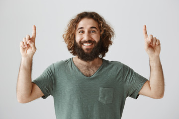 Portrait of thrilled joyful eastern male model with beard and curly hair pointing up with raised hands, smiling cheerfully, being excited while waiting for positive event over gray background