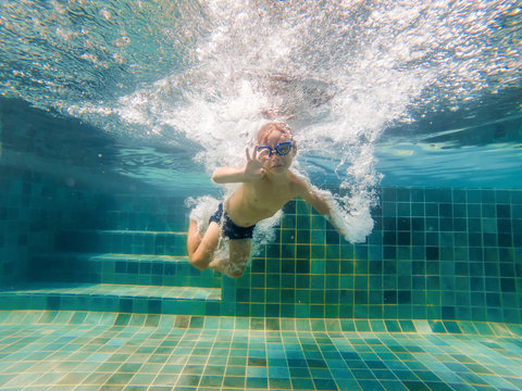 A child boy is swimming underwater in a pool, smiling and holding breath, with swimming glasses