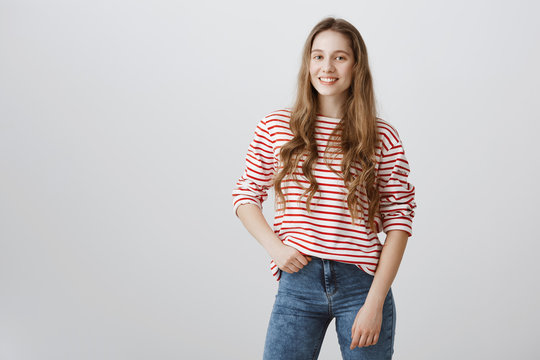 Stylish girl ready to go shopping. Indoor shot of emotive fair-haired teenager in striped sweater holding hand on hip, smiling joyfully, offering her help, wanting to give hand during team work