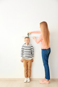 Young woman measuring height of little boy near light wall