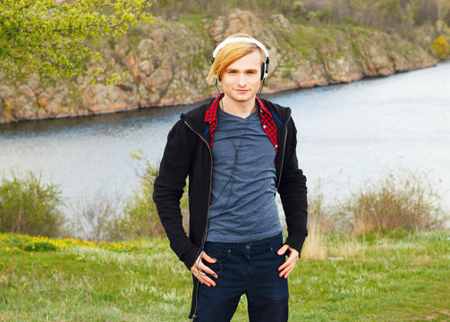 Portrait of young beautiful blond hipster man with headphones standing in nature