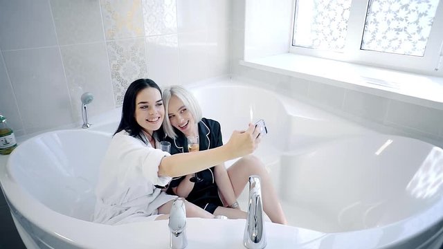 Two sexy women are sitting in empty bathtub and taking selfies on smartphone. They are posing, smiling and laughing in robes.