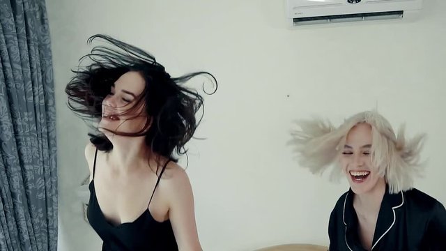 Two pretty and funny girls are jumping in bed together wearing pajamas indoor. They are laughing and screaming.
