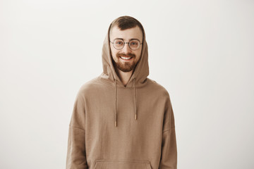 Attractive hipster inviting coworker for cup of coffee. Handsome cheerful european boyfriend in hood and trendy glasses smiling broadly, expressing friendly and positive emotions, confident