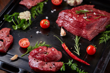 Flat lay of raw beefsteak with vegetables, herbs and spicies on metal tray, close-up, selective focus