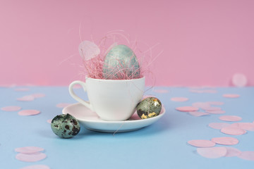 Conceptual art photo of a blue chicken and quail eggs in a white coffee cup on a saucer, decorated with a pink sisal and confetti on a blue and pink background