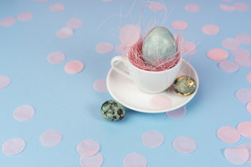 Obraz na płótnie Canvas Мinimalistic conceptual art photo of a blue chicken and quail eggs in a white coffee cup on a saucer, decorated with a pink sisal and confetti on a blue background