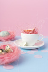 Obraz na płótnie Canvas Yolk of broken egg in shell in a white coffee cup on a saucer, and blue chicken and quail eggs decorated with a pink sisal and confetti on a blue and pink background