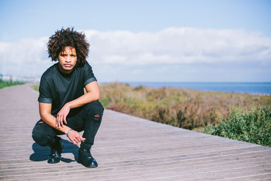 portrait of young man with dark skin, mixed race, big African hair, sitting outdoor on sea shore