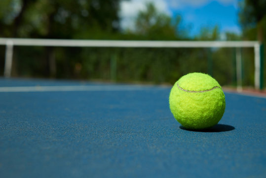 Yellow bright tennis ball is lying on on blue carpet of opened court during sunny day. Made for playing tennis. Contrast image with satureted colors. Concept of tennis outfit photografing.