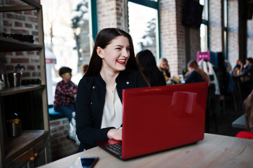 Brunette girl sitting on cafe and working with red laptop.