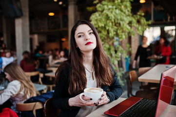 Brunette girl sitting on cafe with cup of cappuccino, working with red laptop.
