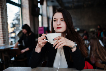 Brunette girl sitting on cafe with cup of cappuccino, listening music on headphones.
