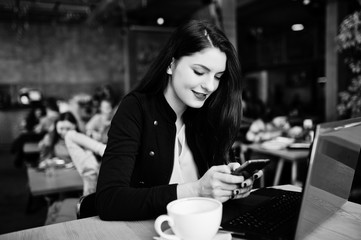 Brunette girl sitting on cafe with cup of cappuccino, working with red laptop and looking at mobile phone.