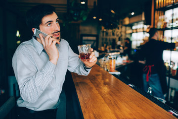 Handsome guy sitting at the barman's stand and talking on the phone. He hold a cup of drink and enjoying his free time in the club.
