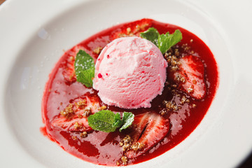 close-up shot of pink ice cream scoop on white plate decorated with red jam, strawberry pieces and mint leaves. ready delicious dessert.