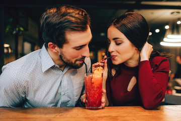 Nice picture of young couple sitting together and drinking red cocktail from the same glass at the...