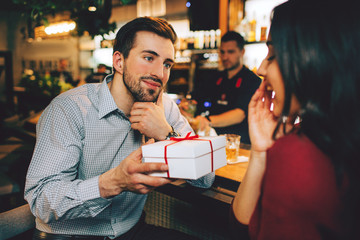 Boyfriend and girlfriend sitting together in restaurant. He has a present for her. She didn't expect that. She looks amazed and happy as so the guy is.