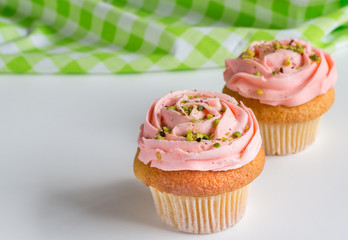 Two pink rose swirled frosted cup cakes with pistachio nut sprinkles on white background