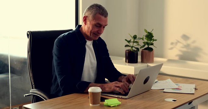 Smiling mature businessman answering his cellphone while sitting at a desk in his office working online with a laptop