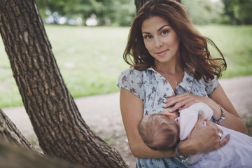 A beautiful smiling young brunette woman with long hair is breastfeeding a pretty baby. They are in the green summer forest. - obrazy, fototapety, plakaty