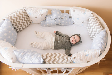 Fototapeta na wymiar Beautiful newborn baby lying in an oval bed with beautiful bumpers in delicate gray, blue, white tones