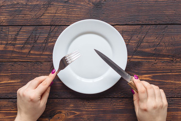 female hands holding fork and knife over the empty plate on dark wooden background