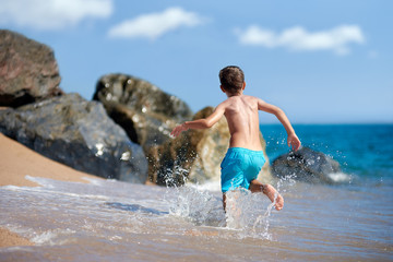 Fototapeta premium Caucasian boy is running in the water along the sea shore. Back view.