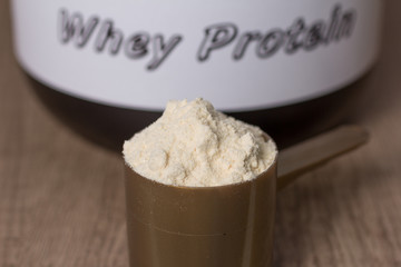 Whey Protein. Golden scoop with vanilla flavour powder and black jar of supplement. Wooden table. Close up, texture.