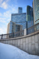 Low angle view of skyscrapers against blue cloudy sky. Snow and iron cast fence in front. End of winter city landscape. Sunset.