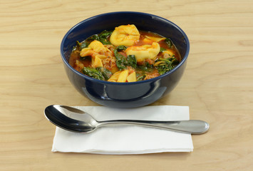 Cheese tortellini tomato stew soup with spinach and sausage in blue bowl with soup spoon and napkin
