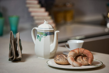Morning breakfast on a ceramic tray in the modern kitchen interior. Kettle, coffee pot, cup of tea, napkins and croissant on kitchen counter