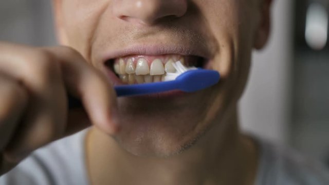 Close-up of young male's mouth as he brushes white natural teeth with blue toothbrush. Toothbrush moving up and down brushing teeth and man smiles to the camera with white toothy smile.
