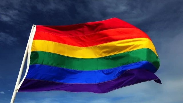 Gay pride rainbow flag flying on a flagpole against bright blue sky in summer sun