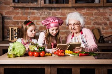 Granny with two granddaughters reading recipe from cooking book and cooking salad