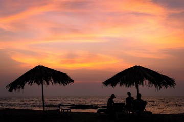 Silhouette of beach umbrellas and people sitting and enjoying golden sunset in Goa, India. Chill out at dusk, summer vacation relax concept. Friends on holidays