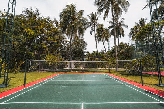 Wide-angle frontal view of a cozy badminton court on a summer day: red and green field with marking on the ground, multiple palm trees, lighting masts on sides