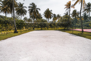Wide-angle frontal view of the volleyball court: coral sand on the ground, multiple palm trees and other plants around, paled tennis court on the right; bright summer day in the Maldives resort