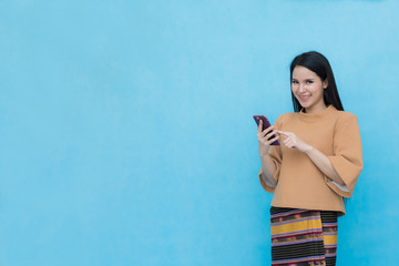 Portrait of sian young girl in traditional thai dress and holding smartphone  isolated on blue sky background. Cotton dress