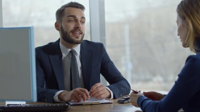 PAN of cheerful salesman in suit sitting at his desk and talking with female customer
