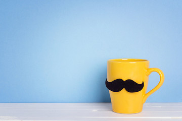 Yellow mug with a mustache on blue background. Copy space. Fathers day holiday.
