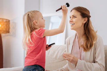 Beauty time. Cheerful pleasant happy girl holding a makeup brush and looking at her mother while having fun with her