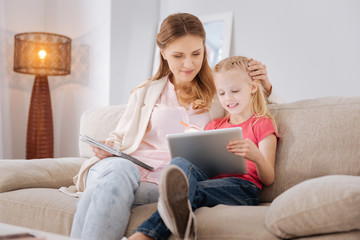 Developing app. Nice smart positive girl sitting together with her mother and using a tablet while playing a developing game