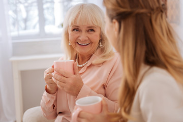Wonderful mood. Happy delighted aged woman smiling and looking at her daughter while having tea...
