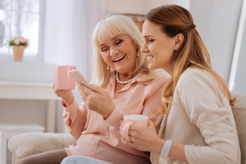 Modern gadget. Happy positive elderly woman having tea and using her smartphone while showing photos to her daughter