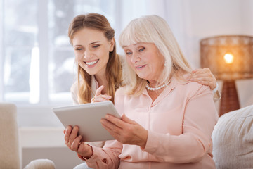 Technological development. Positive nice senior woman smiling and sitting together with her daughter while using new technological device