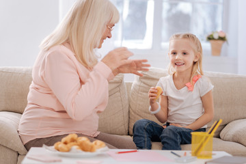 Home made bakery. Positive cheerful pretty girl sitting on the sofa and eating pastry while listening to her grandmother