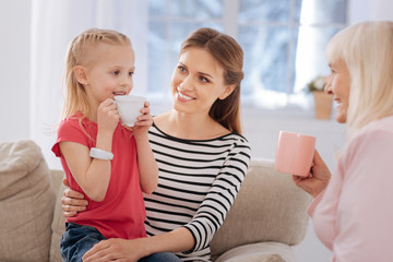 Obraz na płótnie Canvas Favourite drink. Nice joyful cute girl holding a cup with tea and smiling while looking at her grandmother