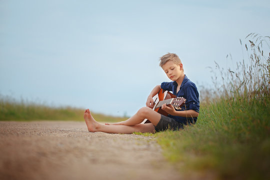 Handsome cute boy is playing on acoustic guitar sittingon road in summer day.